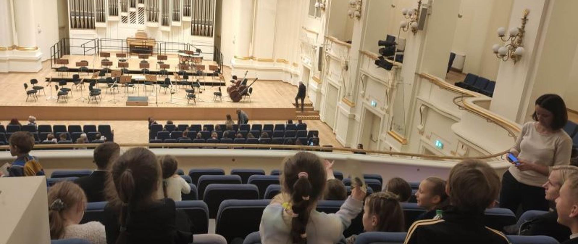 Dzieci w fotelach widowni, koloru szarego, po prawej stronie stoi pani nauczycielka. Tło stanowi scena w kolorze białym z drewnianą podłogą na której stoją krzesła i pulpity na nuty. Po prawej stronie sceny stoją dwa instrumenty.