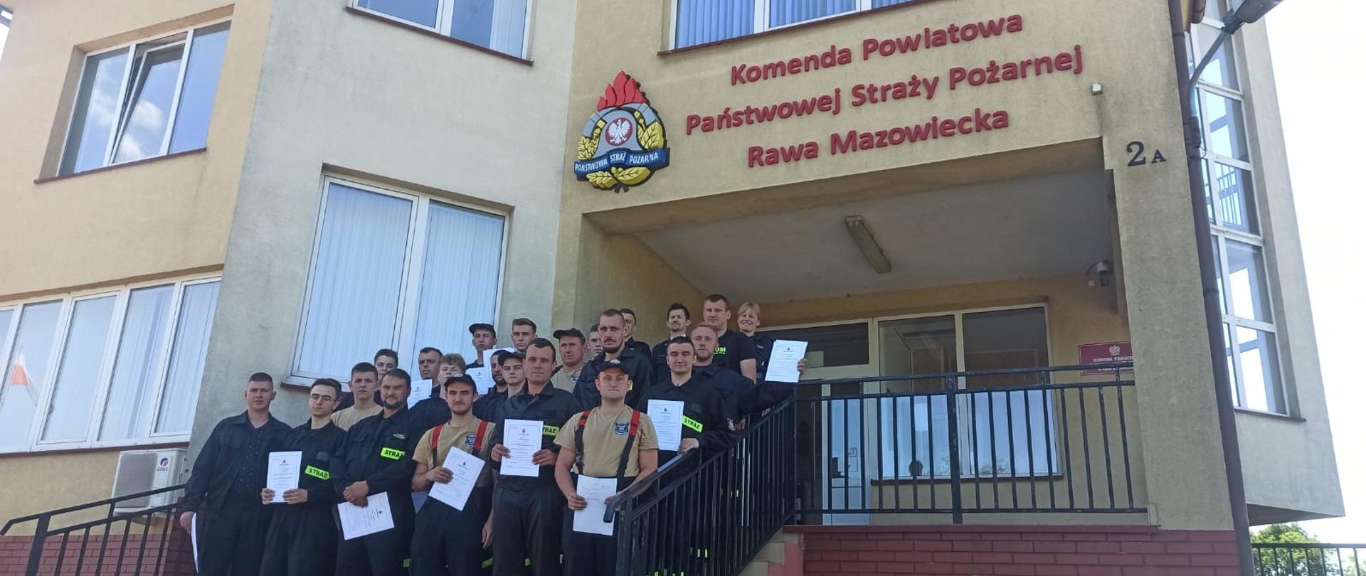 Na schodach zewnętrznych, prowadzących do drzwi wejściowych do komendy, kolejno na stopniach ku górze stoją druhowie OSP, którzy ukończyli szkolenie podstawowe, ubrani w umundurowanie koszarowe, trzymający w dłoniach zaświadczenia o ukończeniu szkolenia. Na samej górze, w ostatnim rzędzie stoi Zastępca Komendanta Powiatowego oraz komisja egzaminacyjna, składająca się z oficerów Komendy Powiatowej Straży Pożarnej w Rawie Mazowieckiej.