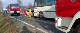 Zdjęcie przedstawia miejsce wypadku. Na pierwszym planie autobus i samochód osobowy. Strażak w ubraniu bojowym wykonuje czynności przy pojeździe osobowym. W tle samochód pożarniczy i odcinek węża leżący na nawierzchni drogi. Na jezdni duża ilość elementów karoserii samochodowej. 