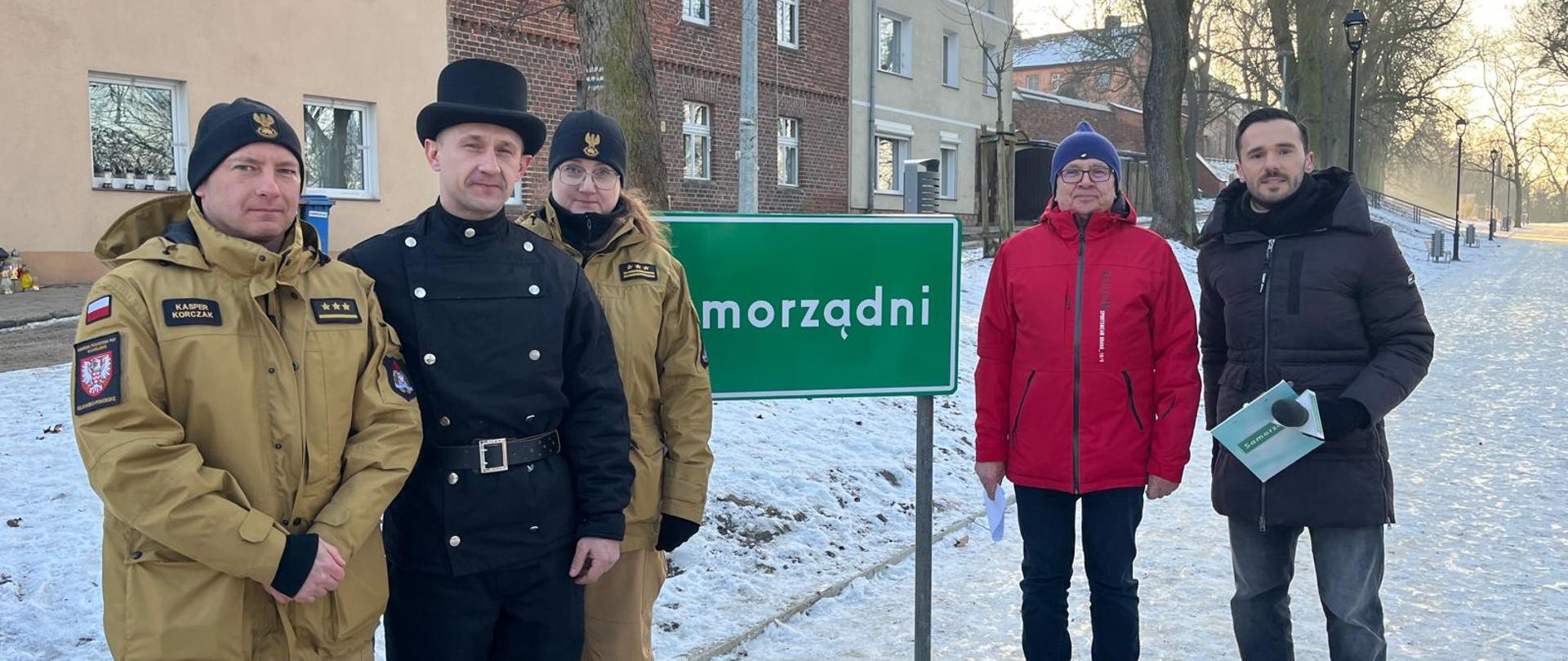 Na chodniku, pokrytym lodem i śniegiem stoi 5 osób, w tym dwoje strażaków w mundurach służbowych, kominiarz w czarnym uniformie, a także dwóch mężczyzn, gdzie jeden w ręku trzyma mikrofon. Pomiędzy nimi tablica na stojaku z napisem Samorządni, na zielonym tle. W dali zabudowa szeregowa domów, drzewa i szereg latarni ulicznych.