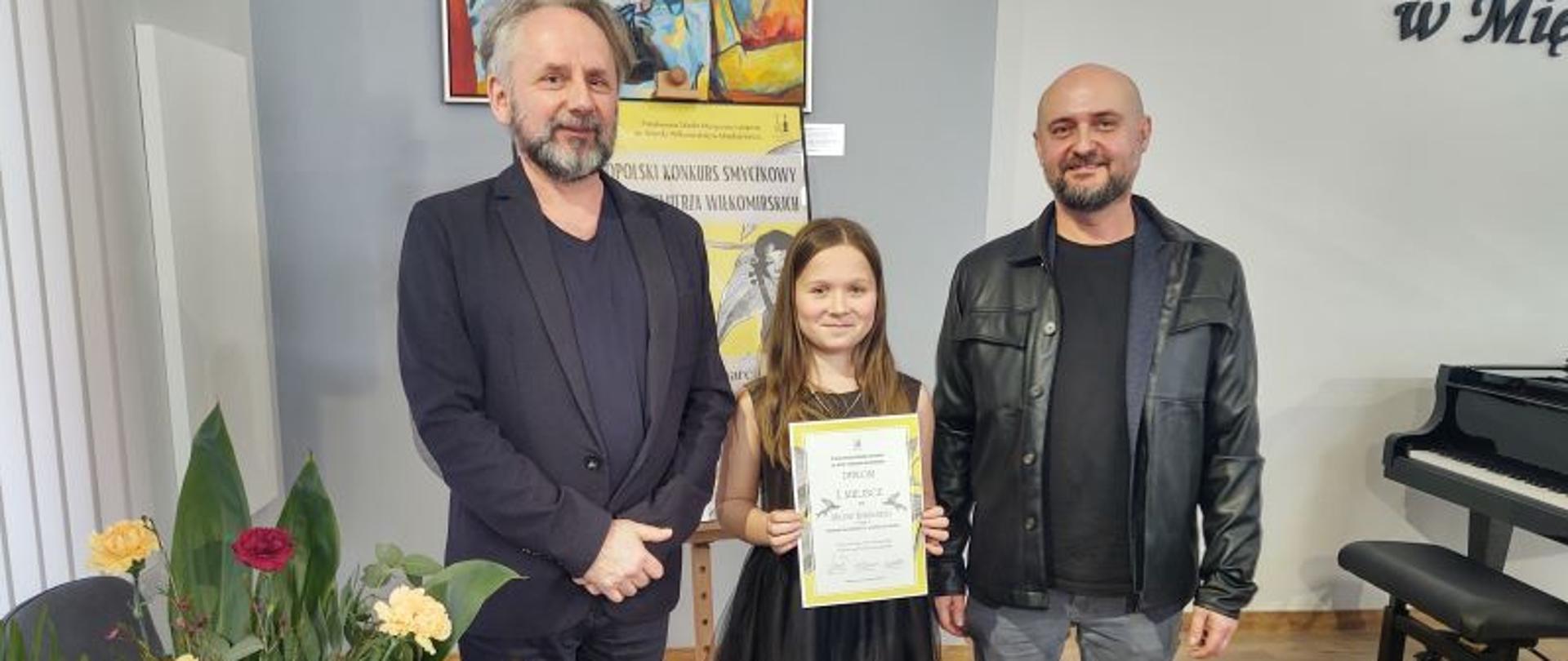 Zdjęcie kolorowe.Od lewej stoją: nauczyciel Tomasz Szymczewski (akompaniator), Milena Kukulska z kl. II c. 6 (ubrana jest w czarną sukienkę i trzyma w rękach dyplom za zajęcie I miejsca w w/w konkursie) oraz Artur Pastuszyński nauczyciel gry na skrzypcach. 