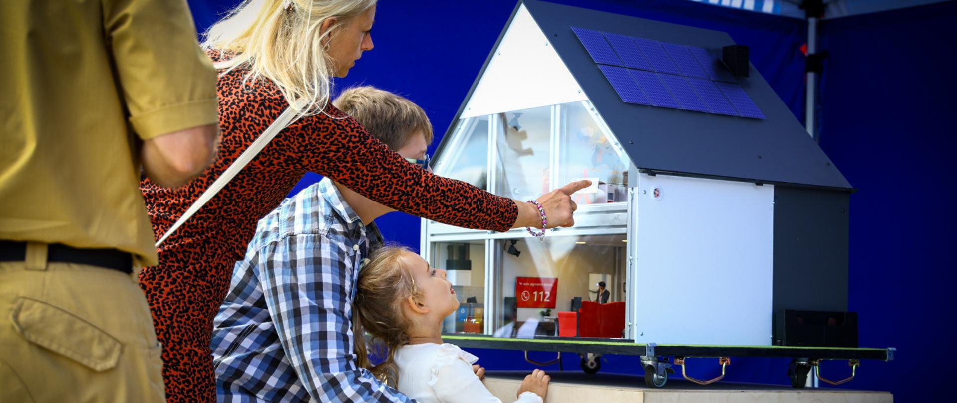 Rodzina z małym dzieckiem ogląda edukacyjną makietę inteligentnego domu z miniaturowym panelem fotowoltaicznym na dachu podczas plenerowego pikniku służb ratunkowych.