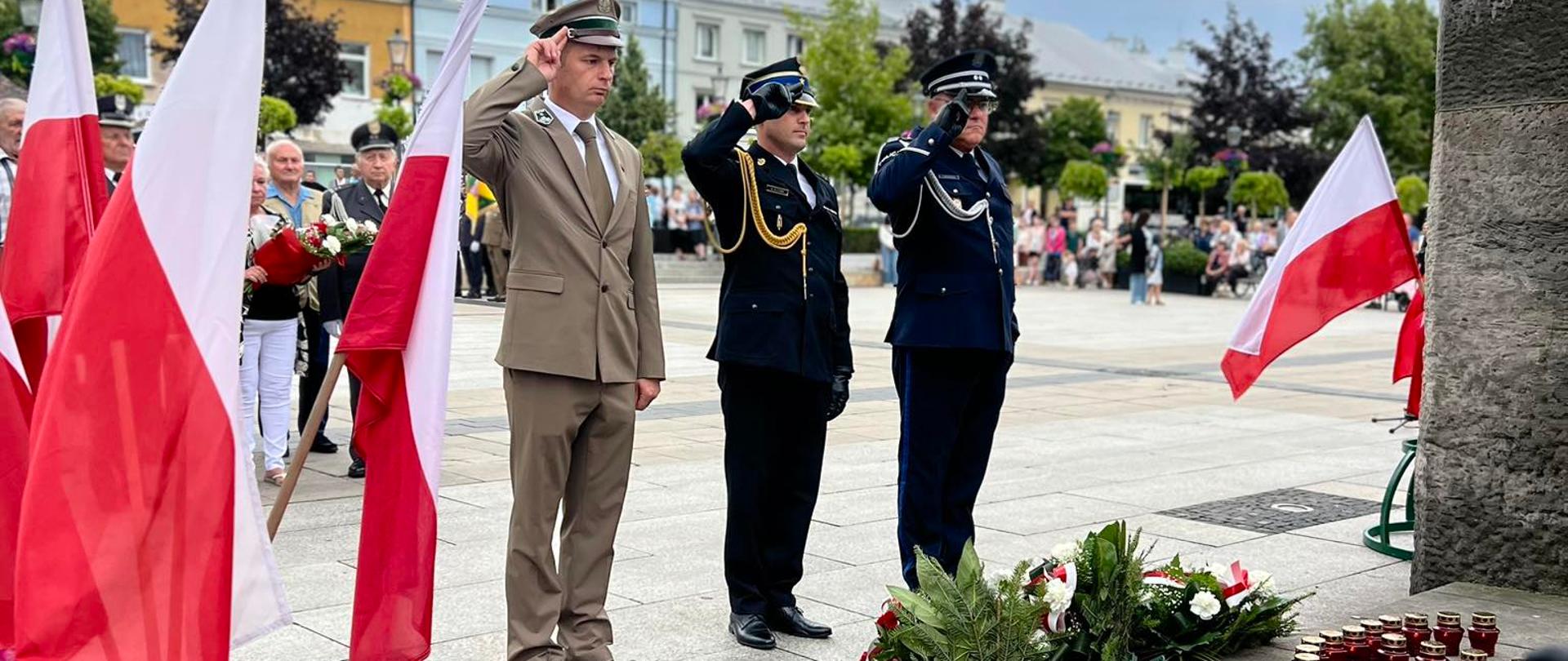 Trzech mężczyzn w mundurach różnych formacji salutuje przed pomnikiem udekorowanym biało-czerwonymi flagami i wieńcami z kwiatów. Dwóch w granatowych mundurach, jeden w mundurze w kolorze khaki. W tle stoją mieszkańcy i kamienice, nad placem zbierają się ciemne chmury. Po środku salutuje Komendant Powiatowy PSP w Grójcu mł. bryg. Marcin Błoński