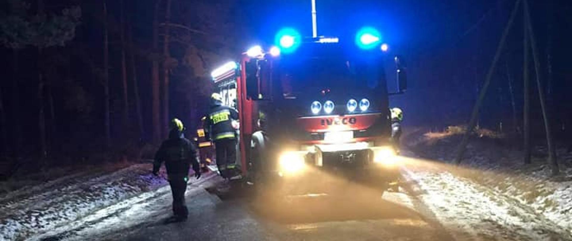 Samochód pożarniczy ustawiony na drodze, noc, dookoła strażacy.