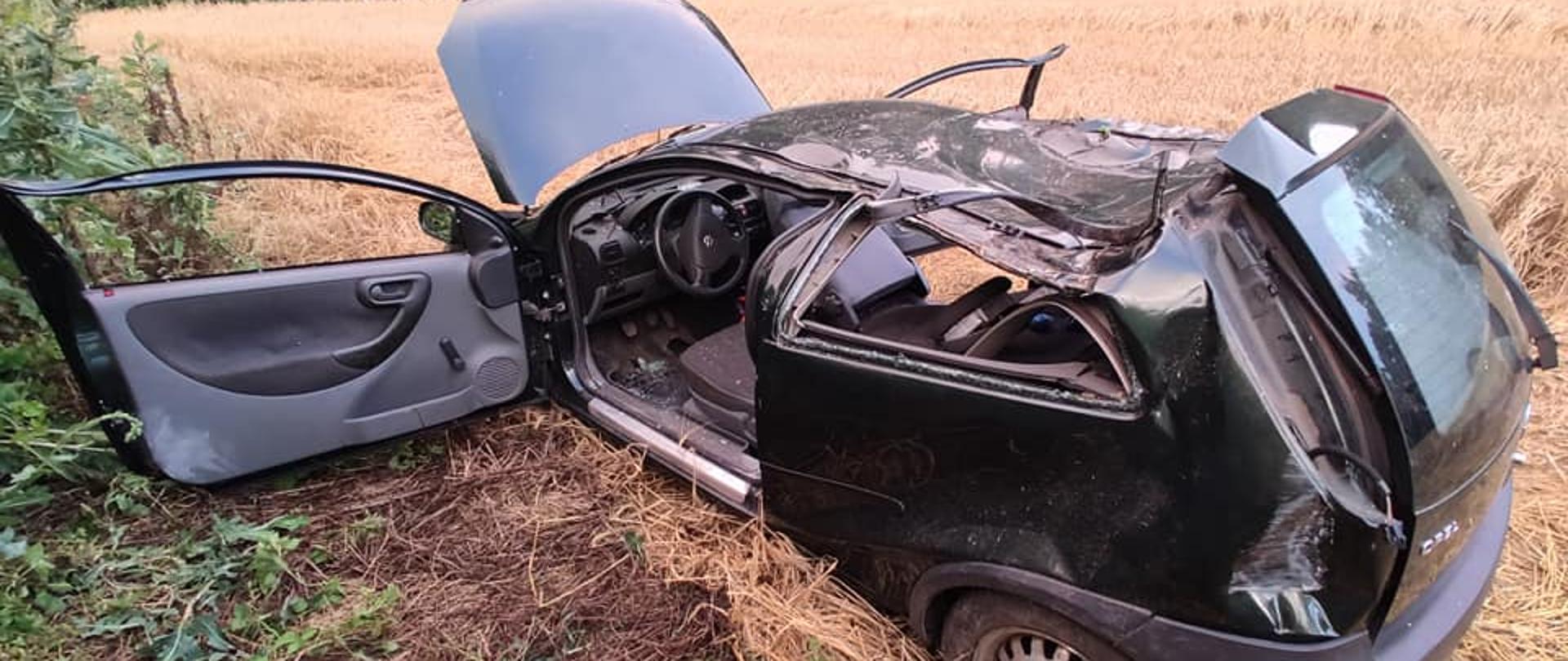 Pole uprawne. W tle wysokie zielone drzewa liściaste. W zbożu stoi na kołach czarny samochód osobowy po wypadku. Otwarta maska i przednie drzwi od kierowcy. Dach i bok pojazdu mocno wgniecione po dachowaniu. 