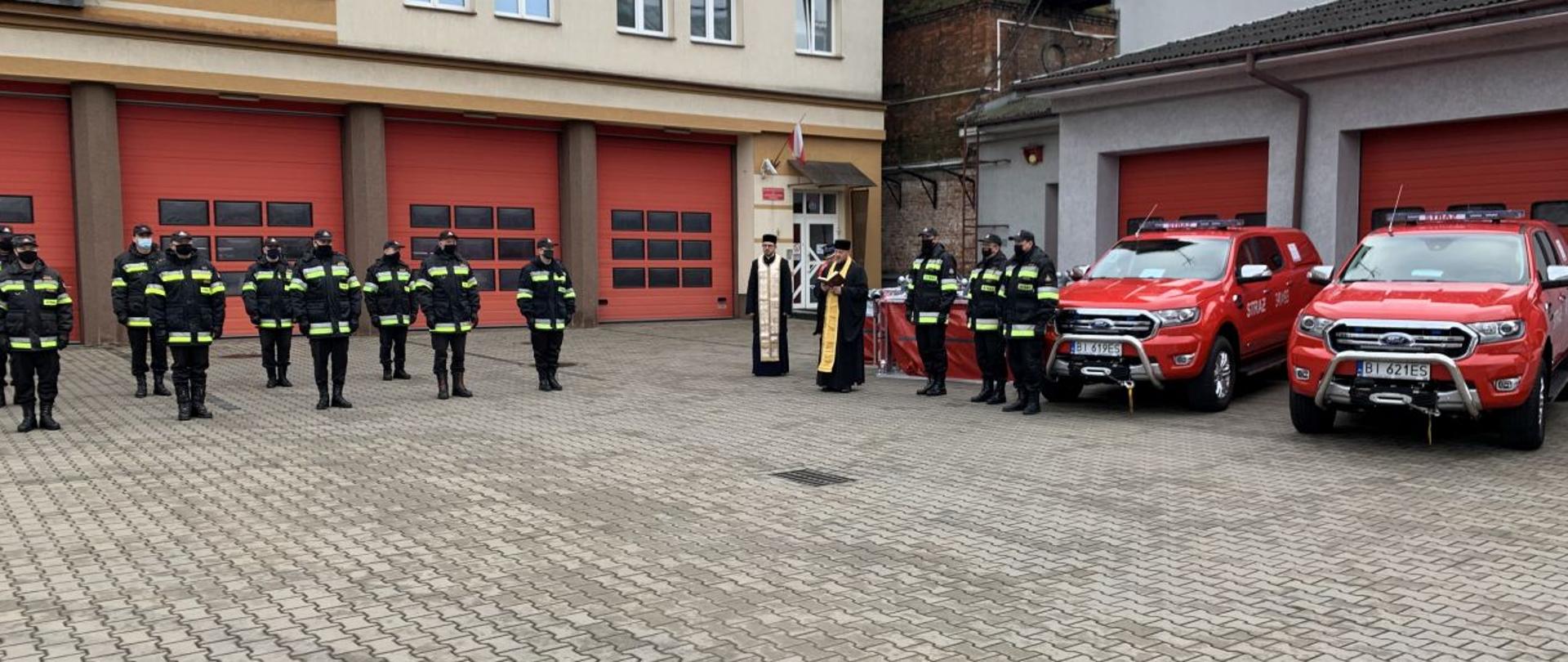 Uroczyste przekazanie pojazdów ratowniczych i sprzętu w KW PSP Białystok.Na zdjęciu widoczni strażacy w umundurowaniu stojący w odstępach zgodnie z reżimem sanitarnym, w tle widoczne garaże JRG 1 w Białymstoku, po prawej stronie widoczny sprzęt przekazywany jednostkom, samochody terenowe i zbiorniki na wodę, na tle pojazdów widoczne kierownictwo komendy wojewódzkiej psp w Białymstoku wraz z kapelanami strażaków.