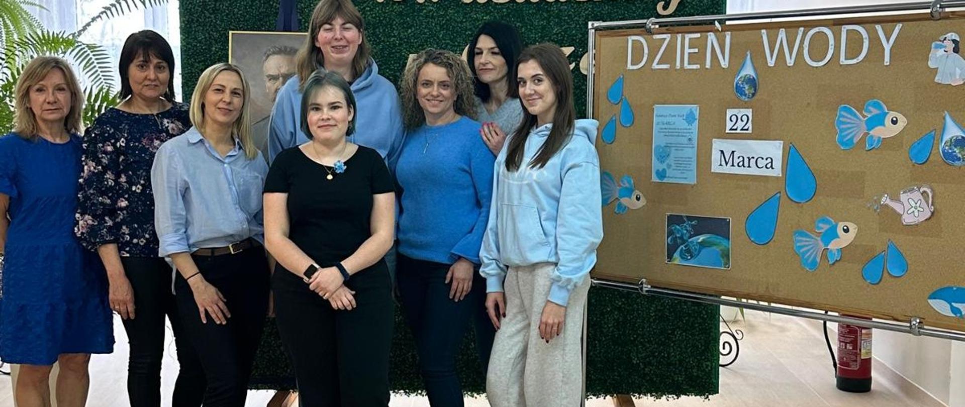 Grupa osób stoi przed zieloną ścianką z nazwą szkoły „ZSCKR im. J. Piłsudskiego”, obok tablicy dekoracyjnej z elementami związanymi z Dniem Wody.
