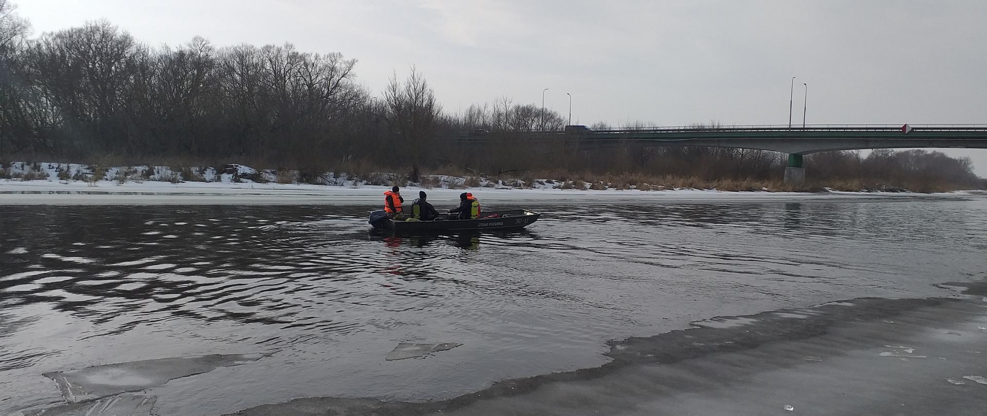 Na zdjęciu widoczna jest oblodzona z dwóch stron brzegów rzeka Narew , środkiem płynie woda. Strażacy w łodzi na środku rzeki płyną w miejsce w którym skoczył z mostu mężczyzna. Na łodzi widać dwóch nurków ubranych w skafandry nurkowe oraz sprzęt. Dwóch strażaków -ratowników ubranych jest w kamizelki ratunkowe . Ratownicy płyną pod most w miejscowości Piątnica. 