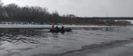 Na zdjęciu widoczna jest oblodzona z dwóch stron brzegów rzeka Narew , środkiem płynie woda. Strażacy w łodzi na środku rzeki płyną w miejsce w którym skoczył z mostu mężczyzna. Na łodzi widać dwóch nurków ubranych w skafandry nurkowe oraz sprzęt. Dwóch strażaków -ratowników ubranych jest w kamizelki ratunkowe . Ratownicy płyną pod most w miejscowości Piątnica. 