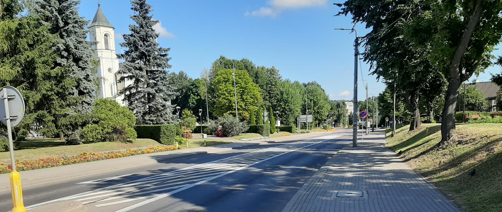 Na pierwszym planie chodnik z polbruku i ulica, w tle po prawej stronie drzewa na trawniku przy chodniku, po lewej stronie ukwiecona rabata, drzewa i za drzewami kościół