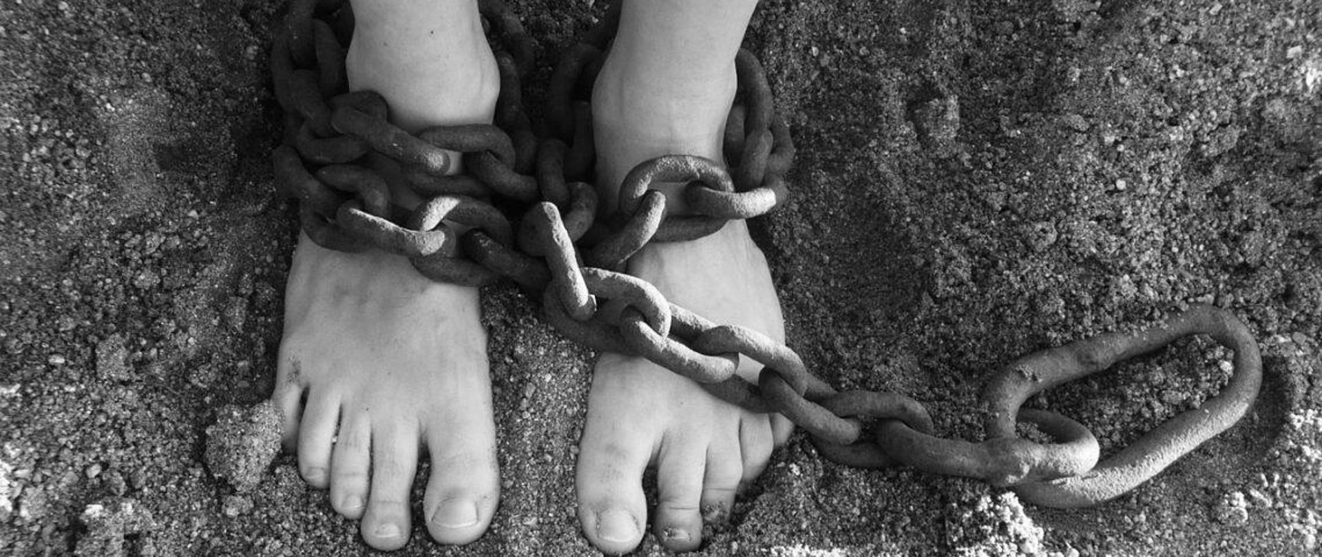 Dwie bose stopy na piasku, związane grubym łańcuchem. Fotografia w biało-czarnym kolorze. 