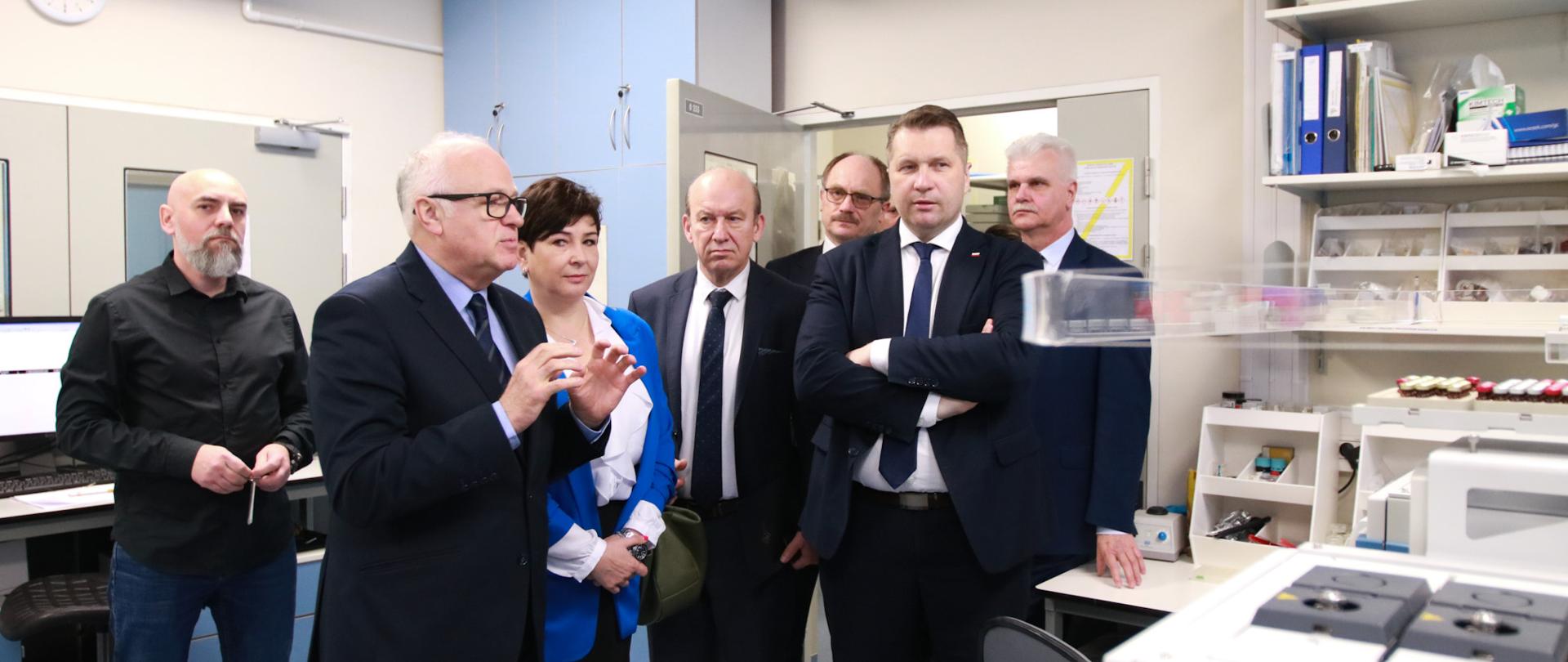 Minister Czarnek w otoczeniu mężczyzn w czarnych garniturach stoi w pomieszczeniu laboratorium, obok niego kobieta w niebieskiej marynarce, pod białymi ścianami stoją różne urządzenia.