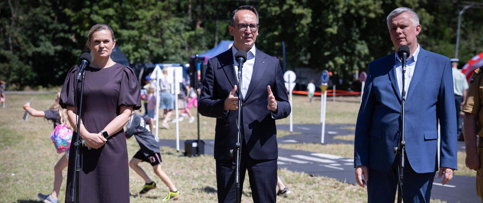 Minister Edukacji Barbara Nowacka, Minister Infrastruktury Dariusz Klimczak oraz Minister Spraw Wewnętrznych Tomasz Siemoniak stoją przy mikrofonach. W tle bawią się dzieci