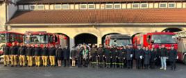 Na tle komendy i pojazdów pożarniczych stoją funkcjonariusze, pracownicy cywilni oraz rodziny strażaków odchodzących na zaopatrzenie emerytalne.