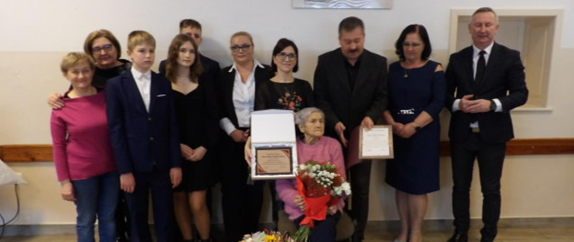 W świetlicy wiejskiej w Prusach na tle ściany z wiszącą grafiką pozuje do zdjęcia 10 osób, w tym dwóch chłopców i dziewczynka. Przed nimi na krześle siedzi starsza kobieta z bukietem kwiatów i dyplomem w ręce, obok jej nóg stoi kosz z kwiatami. 