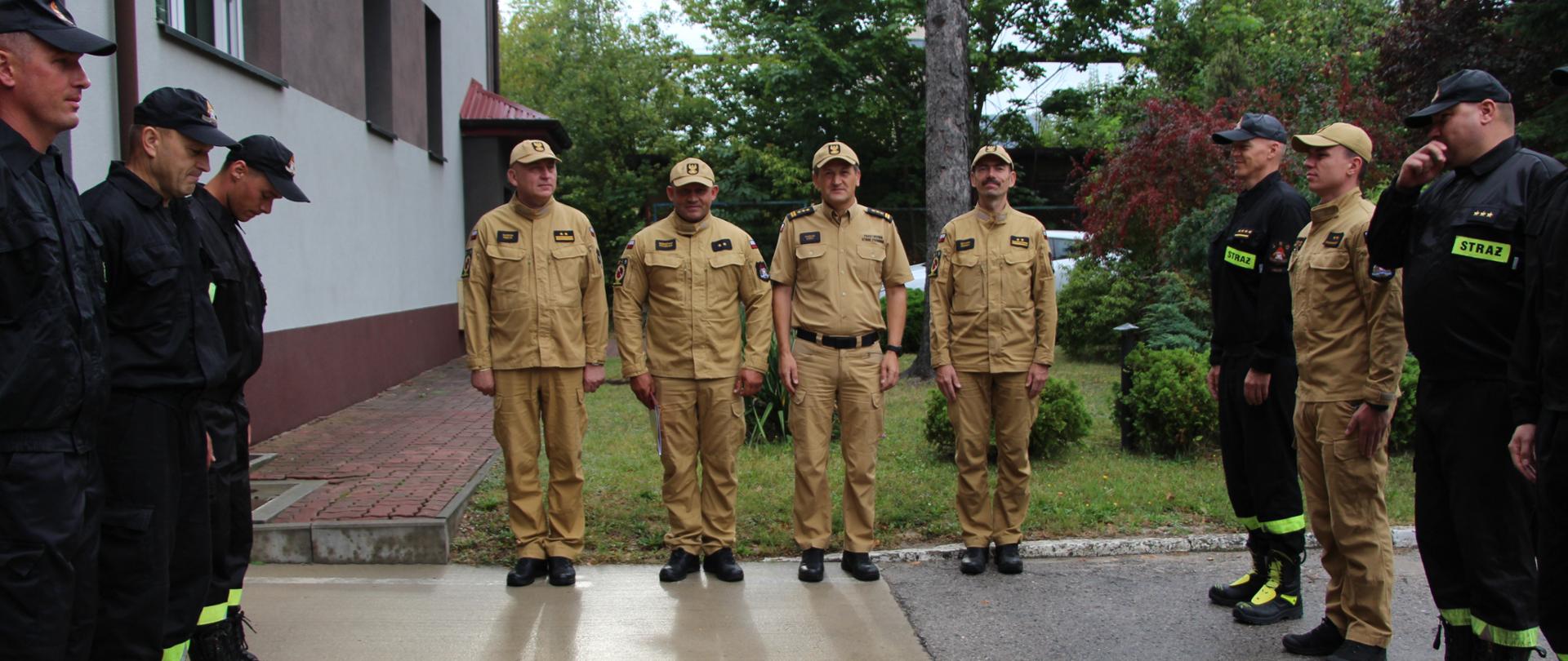 Zdjęcie przedstawia uroczystą zbiórkę przy budynku Jednostki Ratowniczo-Gaśniczej numer 2 w Kielcach. W dwóch szeregach stoją zmiany służbowe, a w centralnym punkcie stoją: komendant miejski, dowódcy jednostki i wyróżniony strażak.