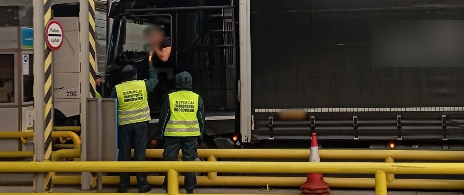 Inspektorzy lubuskiej Inspekcji Transportu Drogowego stoją obok kabiny ciężarówki zatrzymanej przed bramkami poboru opłat na autostradzie A2 i kontrolują trzeźwość kierowcy.