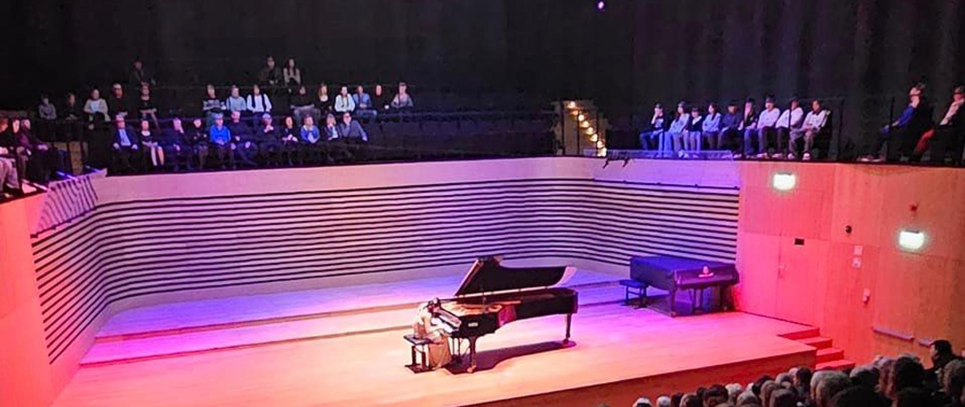 Wnętrze sali koncertowej wypełnione widzami. Na scenie pianistka grająca na fortepianie