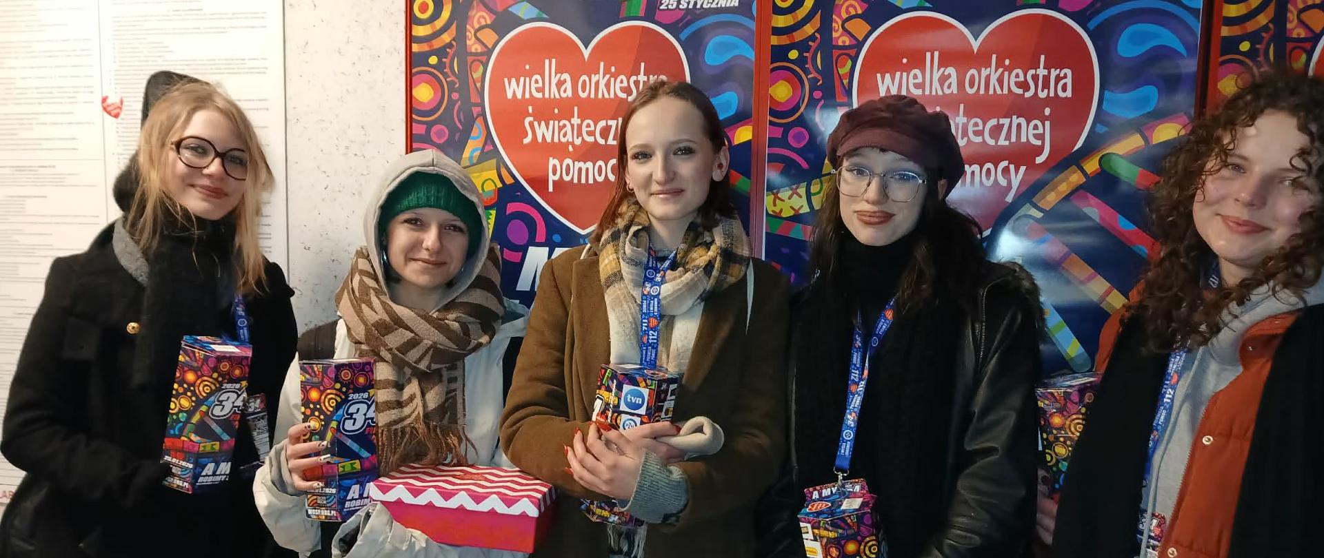 Na pierwszym planie stoi pięć dziewczyn w zimowych kurtkach, w rekach trzymają kartonowe puszki Wielkiej Orkiestry Świątecznej pomocy. Na szyjach maja identyfikatory wolontariuszy 34 finału WOŚP. Na ścianie za ich plecami widoczne są trzy , kolorowe plakaty z czerwonymi sercami w centrum z napisem Wielka Orkiestra Świątecznej Pomocy
