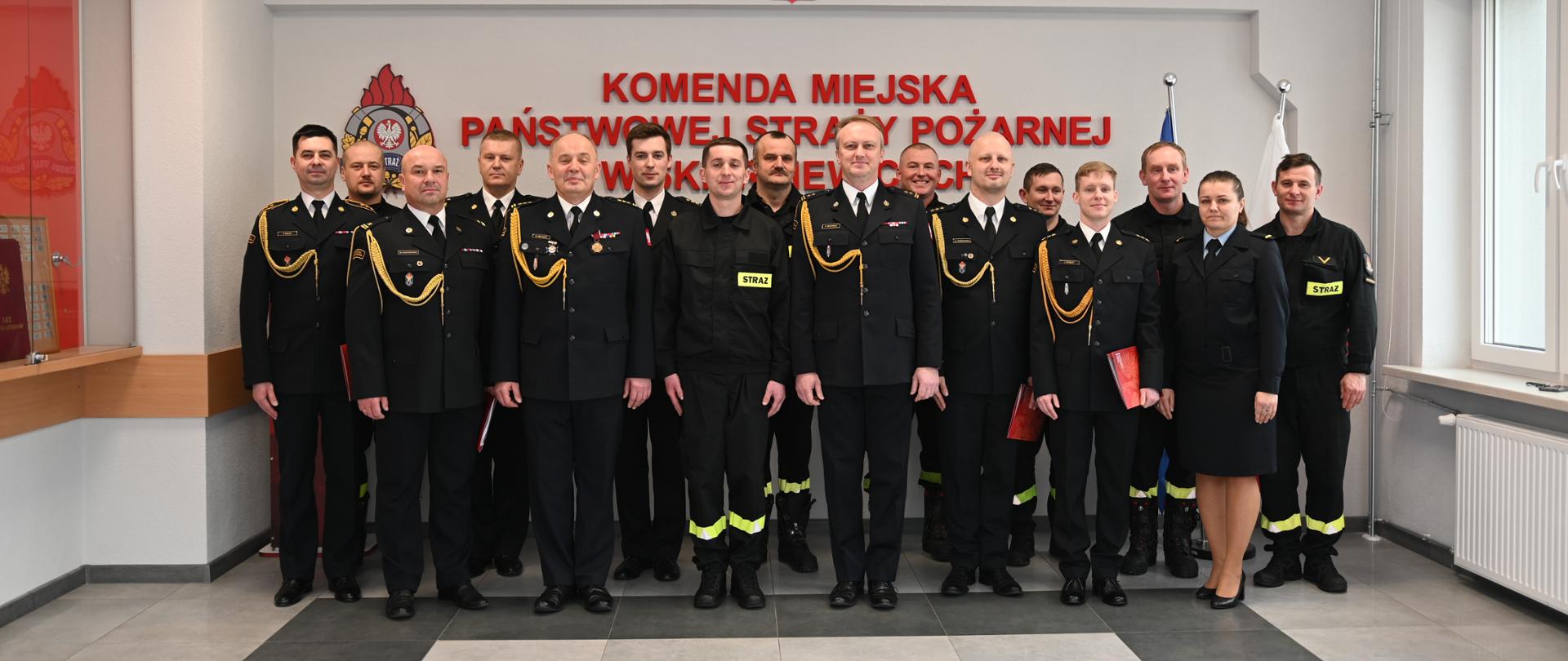 Strażacy w mundurach koloru czarnego na tle i ściany z napisem koloru czerwonego.