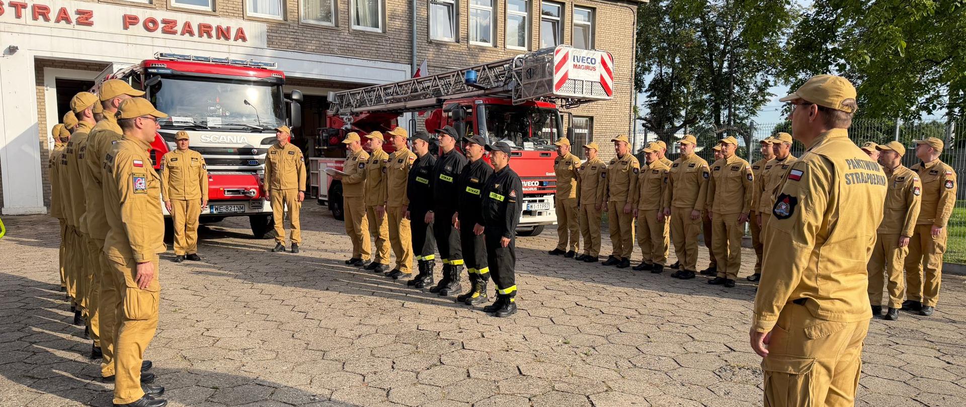 Na placu przed budynkiem stoi grupa strażaków w piaskowych mundurach, ustawiona naprzeciw komendanta. W tle znajdują się czerwone wozy strażackie, w tym samochód z wysuwaną drabiną, a po prawej stronie drzewa i ogrodzenie. Na pierwszym planie widoczny jest zwinięty żółty wąż strażacki.