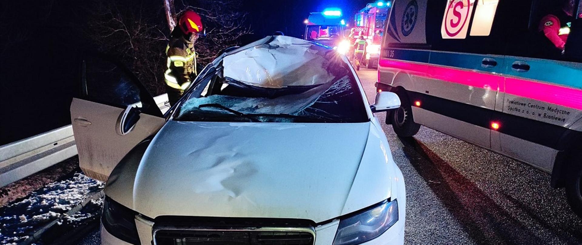 Zdjęcie zrobione nocą. Na zdjęciu białe auto z rozbitym szybą i pogiętym dachem, przy aucie z lewej strony strażak. Z prawej strony karetka pogotowia a w oddali samochody strażackie.