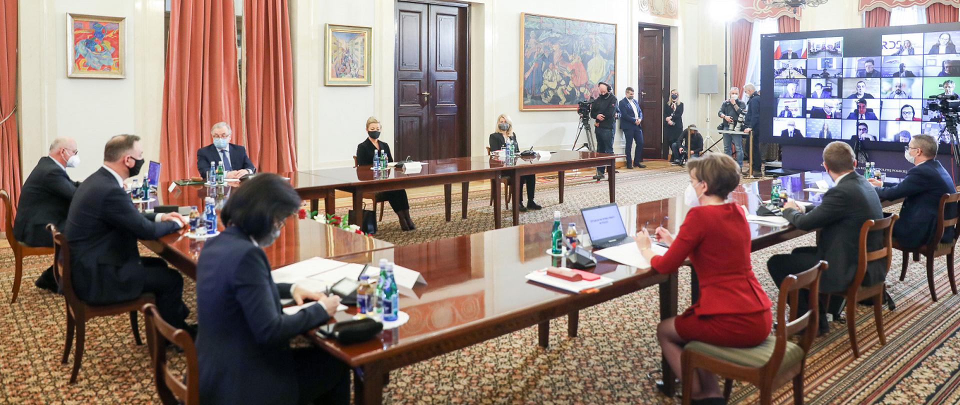 W sali przy stole ustawionym w podkowę siedzi grupa osób. Przed nimi duży monitor, na którym widać zdalne łączenie.