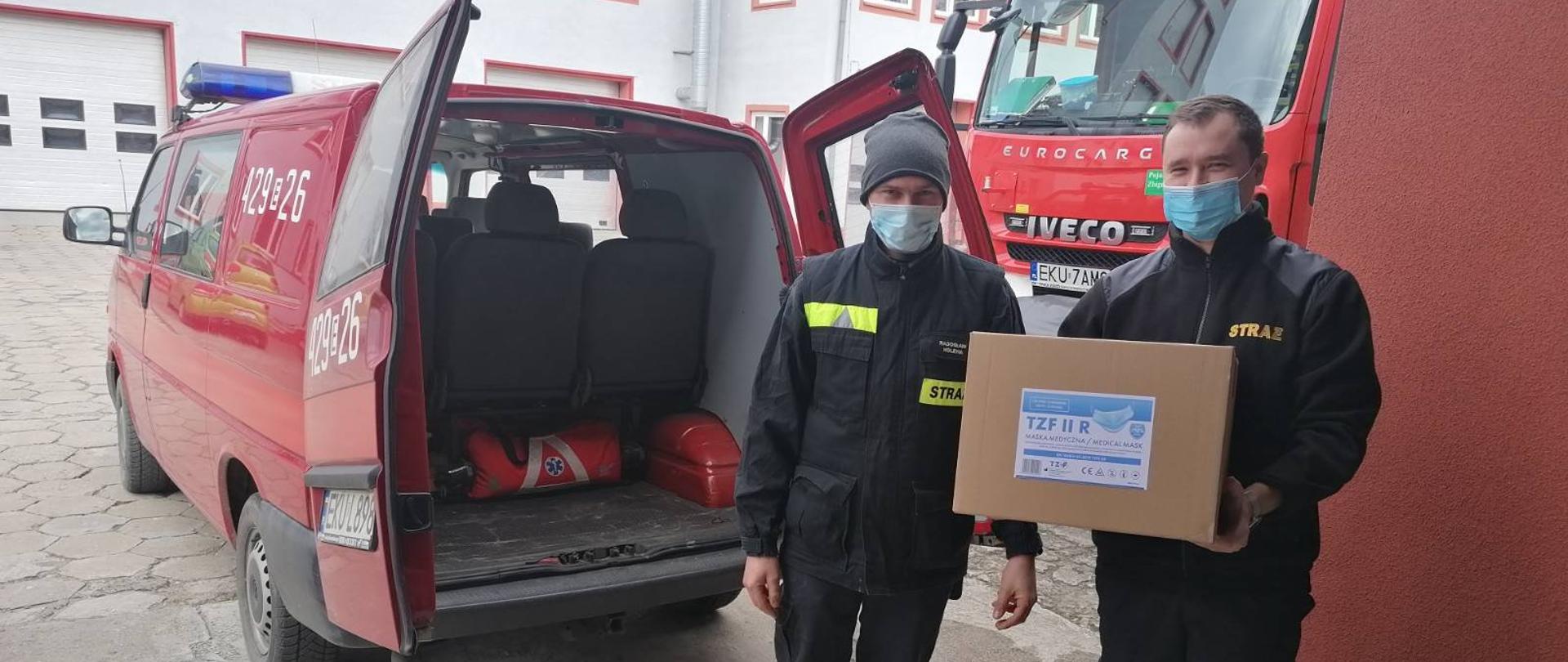 Przed garażem Jednostki Ratowniczo-Gaśniczej stoi dwóch strażaków w mundurach koszarowych koloru czarnego. jeden z nich trzyma opakowanie ze środkami dezynfekcyjnymi. Tyłem do garażu zaparkowany jest samochód strażacki typu bus z otwartymi tylnymi drzwiami.