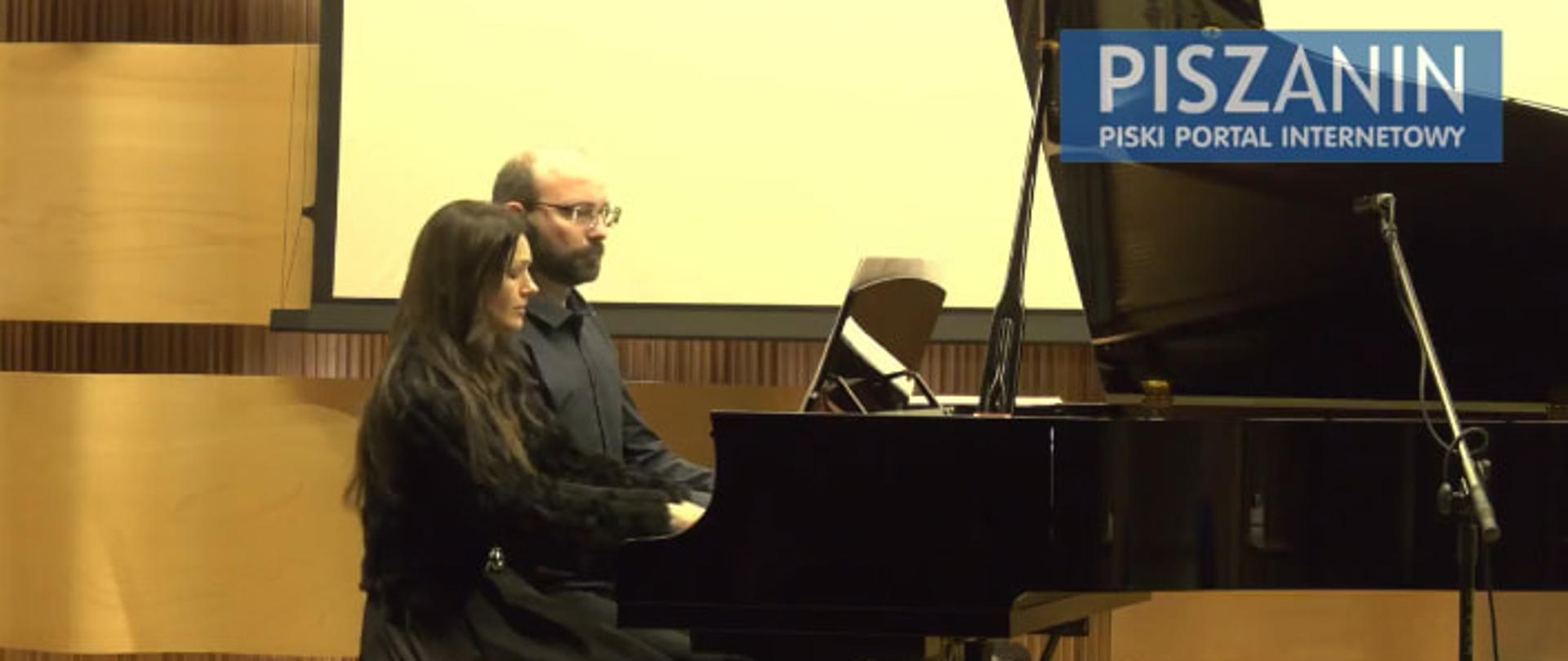 Na tle paneli akustycznych oraz ekranu do projektora dwóch pianistów gra na cztery ręce przy otwartym fortepianie.