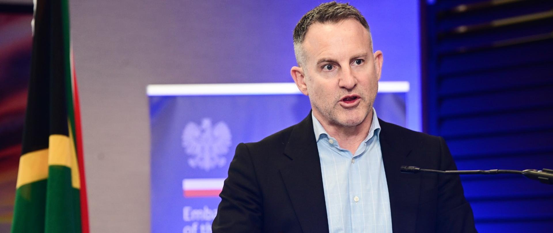 Juliusz Gojło, reprezentujący Ministerstwo Rozwoju i Technologii RP przemawia przy mównicy; z lewej strony flaga RPA; w tle roll-up Ambasady RP w Pretorii