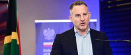 Juliusz Gojło, reprezentujący Ministerstwo Rozwoju i Technologii RP przemawia przy mównicy; z lewej strony flaga RPA; w tle roll-up Ambasady RP w Pretorii