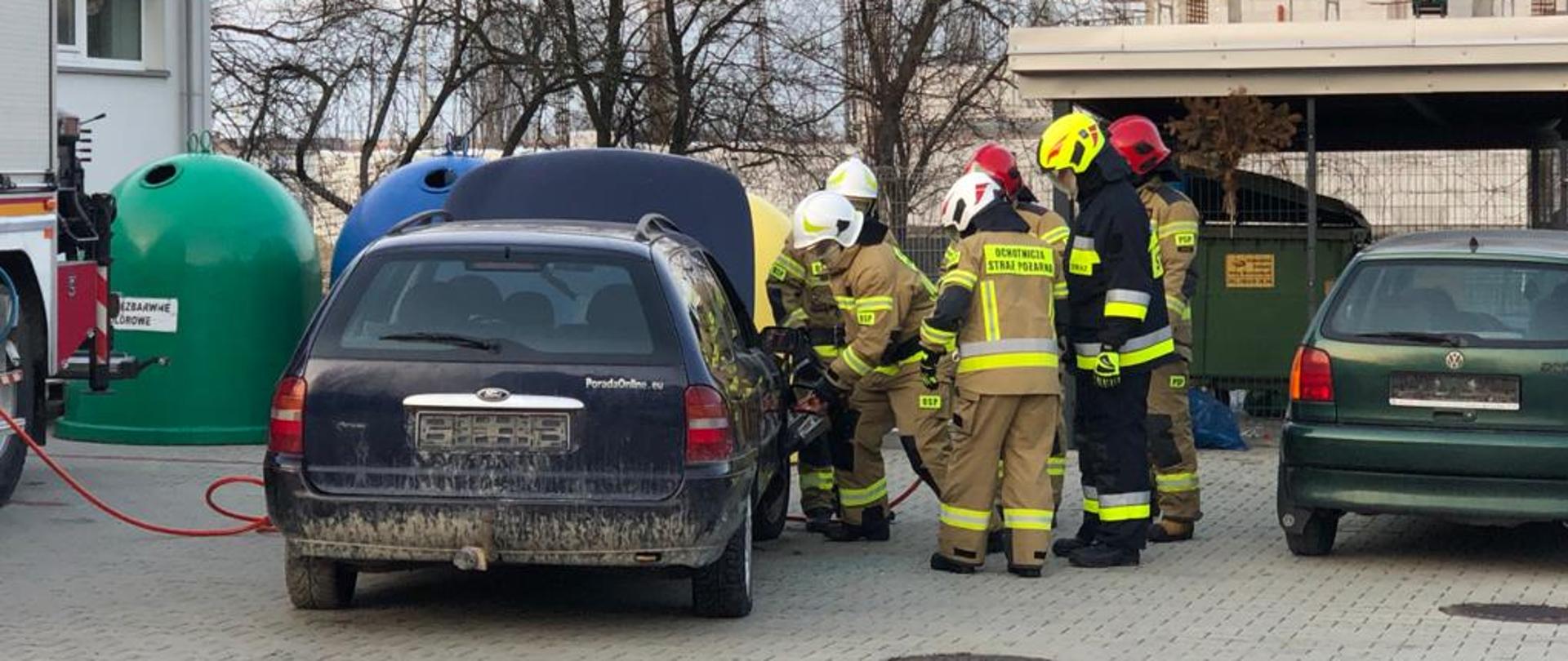 Grupa 4 strażaków z Ochotniczej Straży Pożarnej ubranych w umundurowanie specjalne koloru piaskowego używając specjalistycznego sprzętu hydraulicznego próbuje wywarzyć przednie prawe drzwi do samochodu marki Ford Mondeo Kombi koloru niebieskiego. Dwóch strażaków Komendy Powiatowej PSP w Bochni nadzoruje zadane ćwiczenie w ramach egzaminu. Obok stoi drugi wrak samochodu marki Volkswagen koloru zielonego. W pobliżu znajduje się również duży samochód strażacki. Całe ćwiczenie odbywa się na placu wewnętrznym Komendy PSP w Bochni. 