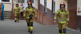 Ćwiczenia w ZSZ. Z jednego z budynków szkolnych wychodzi trzech strażaków, którzy nadzorowali przebieg ewakuacji. Maja na sobie ubrania specjalne koloru piaskowego oraz czerwone hełmy. 
