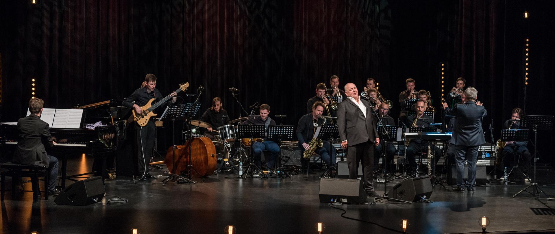 Zdjęcie zespołu jazzowego na scenie, dyrygent stoi tyłem, wokalista z przodu. 