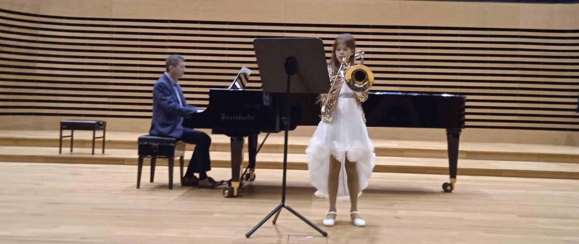 Uczennica stoi przed fortepianem i gra na puzonie, nauczyciel gra na fortepianie na scenie sali koncertowej PSM