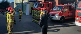 dwunastu strażaków stoi przed budynkiem komendy na baczność w dwóch rzędach naprzeciw siebie . Z lewej strony przodem do nich stoi komendant powiatowy