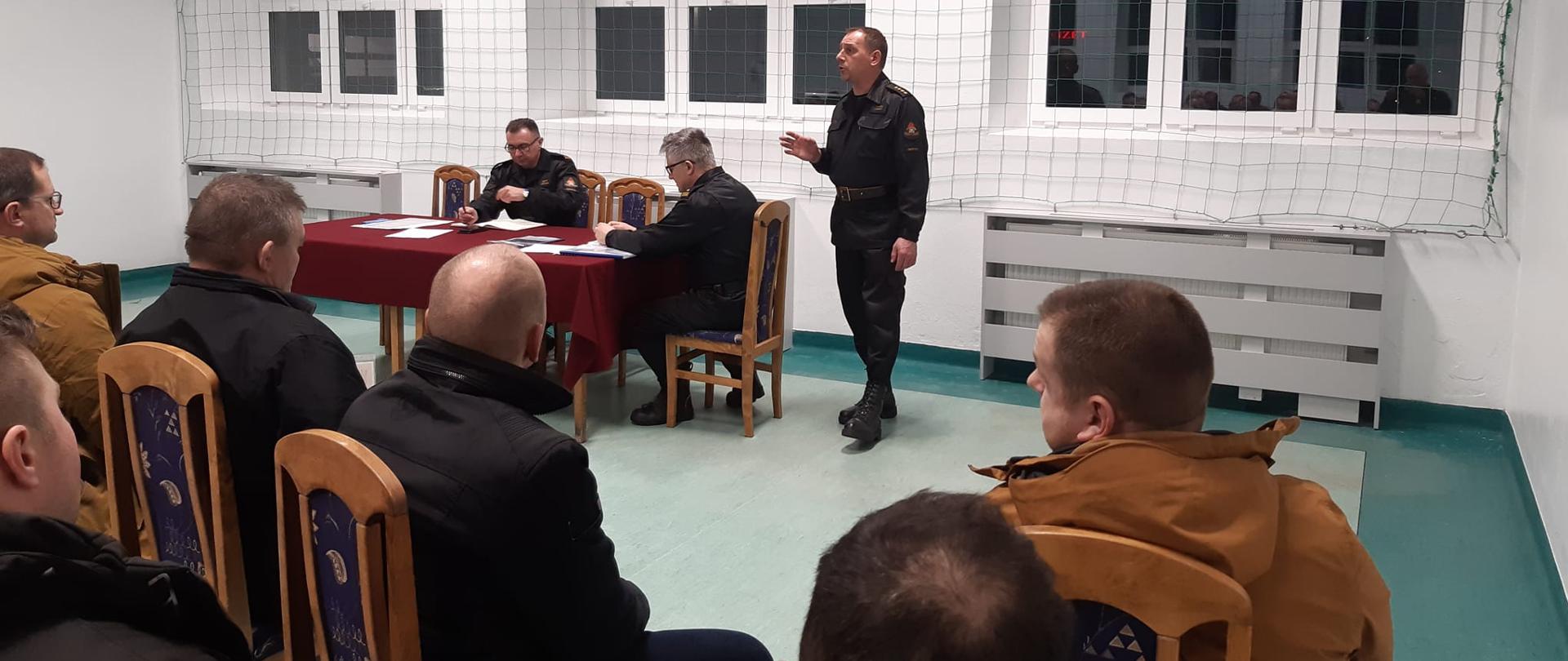 Na zdjęciu widać dziewięciu mężczyzn. Jeden stoi a reszta siedzi. Dwóch mężczyzn ubranych jest w brązowe kurtki a reszta w czarne stroje. Na stole rozłożone jest czerwone sukno, na którym leżą dokumenty. W tle widać okna. Cała ściana przesłonięta jest zieloną siatką. Za oknami widać, że jest ciemno. Zdjęcie zdobione jest w środku pomieszczenia, gdzie zapalone jest światło.