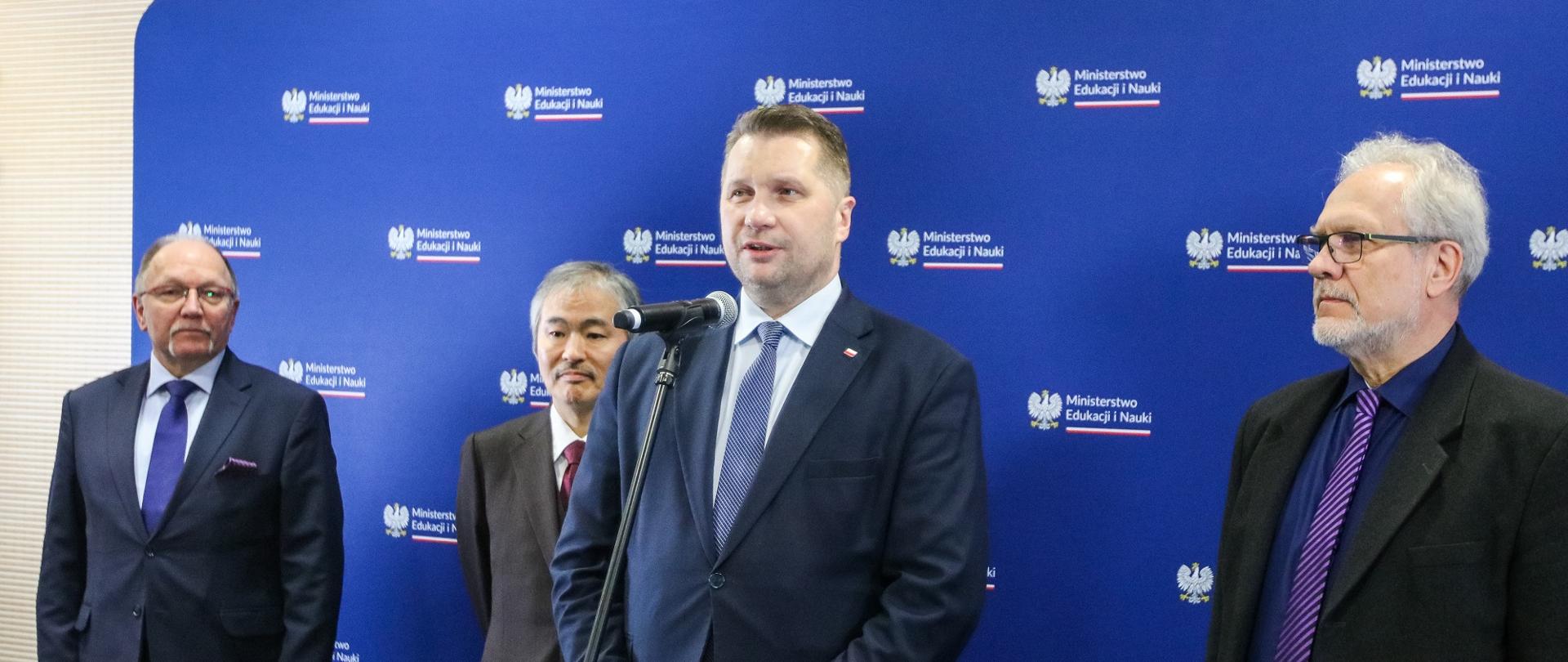 Minister Czarnek stoi przed niebieską ścianka i mówi do mikrofonu na stojaku, obok niego trzech mężczyzn w garniturach.