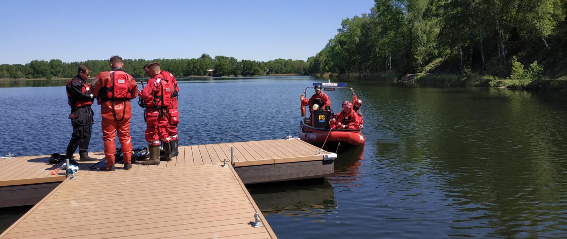 Czterech strażaków ubranych w ubrania specjalne ratowników wodnych stoi na pomoście, czterech innych podpływa do pomostu na pontonie