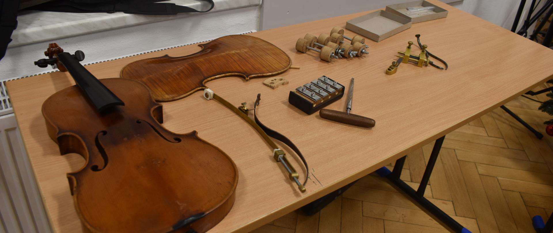 Na zdjęciu, na ławce szkolnej widnieją rozklejone skrzypce oraz narzędzia lutnicze służące do renowacji skrzypiec. Kolorystyka zdjęcia jest brązowo-szara.