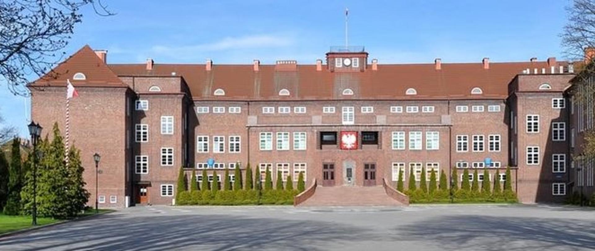Szkoła Policji w Słupsku: budynek z czerwonej cegły, piętrowy, nad głównym wejściem godło Polski, wokół zieleń. 