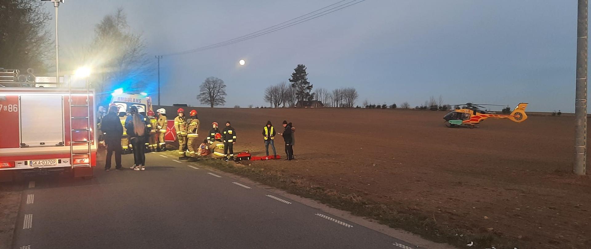 Teren działań przy potrąceniu grupy osób. Z lewej strony tyło samochodu strażackiego z wysuniętym masztem oświetlającym miejsce zdarzenia. Obok po prawej usytuowana karetka pogotowia przed którą na poboczu znajdują się strażacy udzielający pomocy osobom poszkodowanym i prowadzący czynności funkcjonariusze policji. Z prawej strony na polu znajduje się śmigłowiec LPR. 