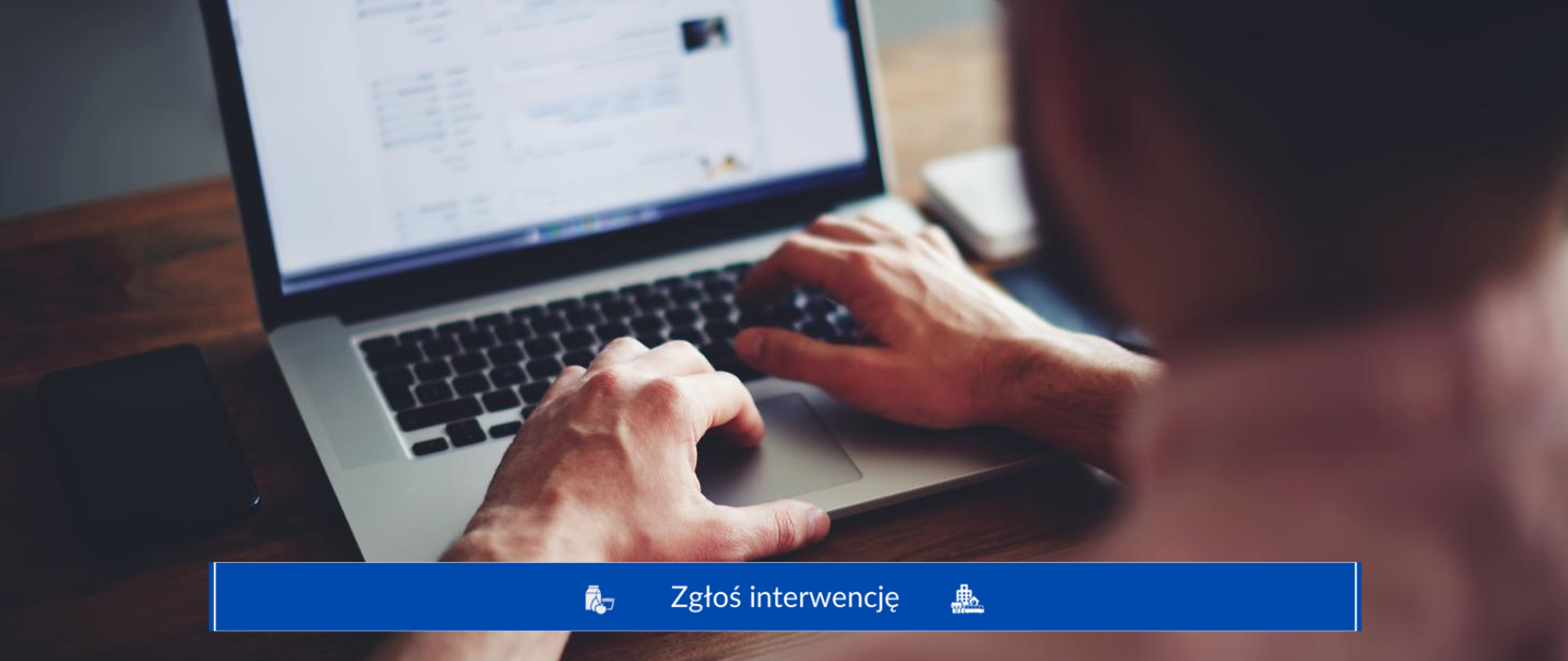 Obraz ukazuje osobę siedzącą przy stole z laptopem, z rękami na klawiaturze. Na dole zdjęcia znajduje się niebieski pasek z tekstem "Zgłos interwencję", wskazującymi na opcję zgłaszania interwencji. Tło jest rozmyte, skupiając uwagę na dłoniach i laptopie.