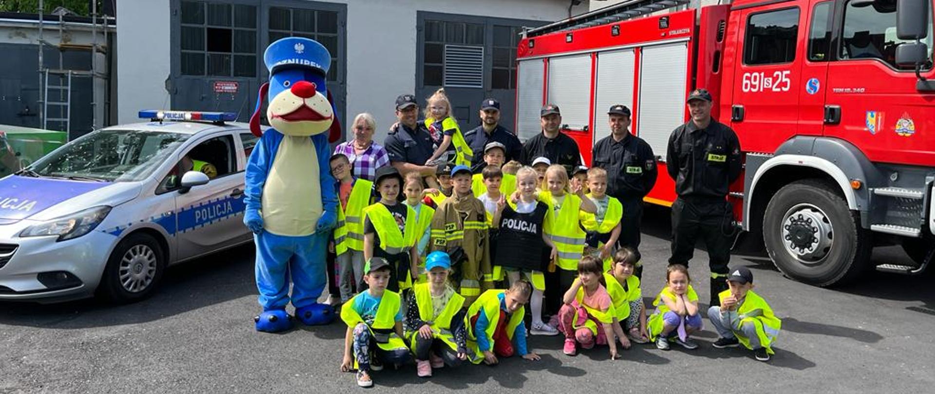 Dzieci, strażacy, policjanci i policyjny sznupek stoją na tle samochodu pożarniczego i radiowozu. W tle budynek Komendy Policji.