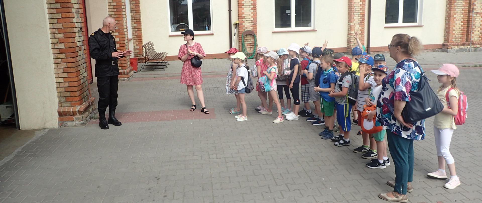 Funkcjonariusz PSP stojący przed garażami a z nim grupa dzieci z opiekunkami.