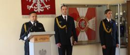 Na zdjęciu strażacy stoją w mundurach wyjściowych, jedna osoba przemawia za mównicą, na ścianie wisi godło Polski i sztandar.
