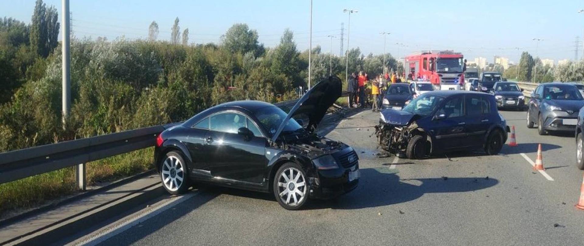 Na zdjęciu, na pierwszym planie widoczny jest rozbityb samochód marki Audi. Za nim rozbity samochód marki Renault. Na druugim planie wóz strażacki i ratownicy.
