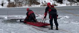Zdjęcie przedstawia przemyskich strażaków podczas ćwiczenia z zakresu ratownictwa wodno-lodowego , trzech strażaków w skafandrach suchych i kaskach ochronnych w trakcie podejmowania z przerębla strażaka-pozoranta na sanie lodowe