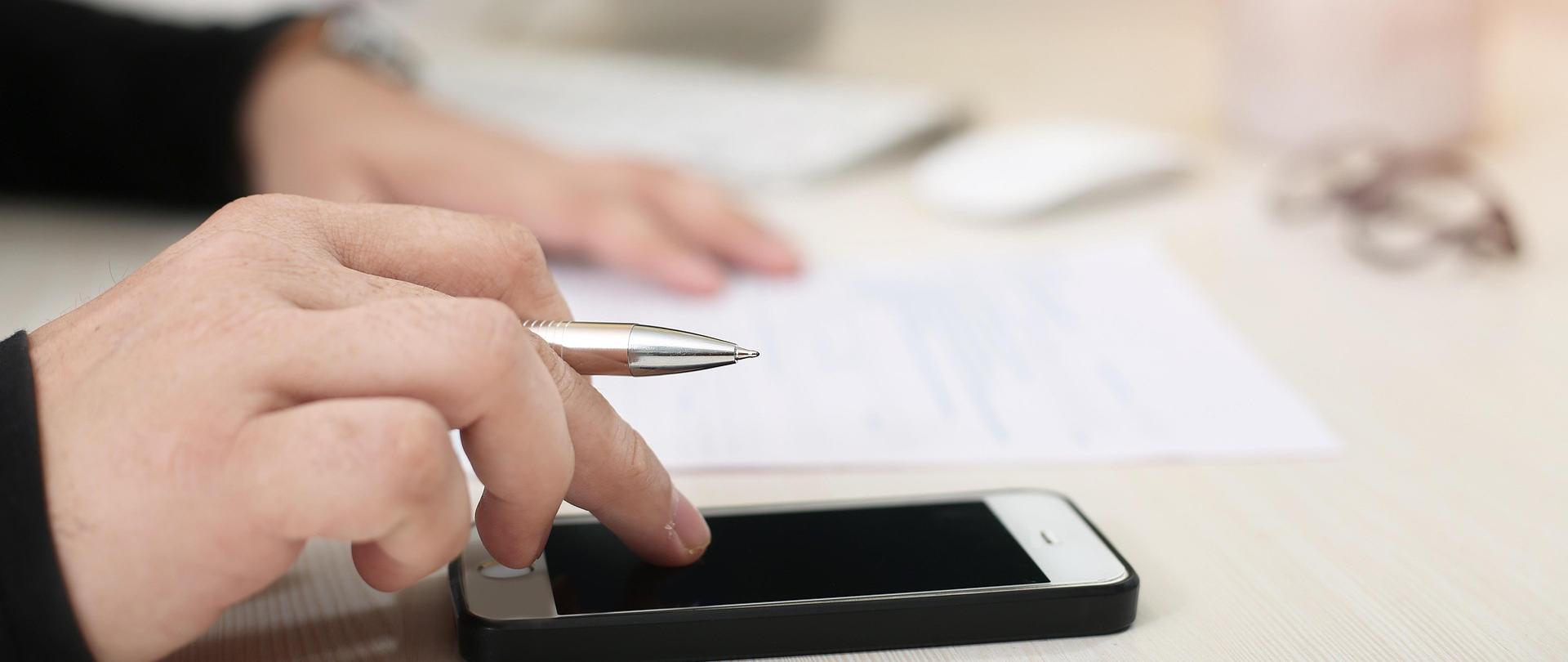 Zdjęcie przedstawia dłonie oparte na biurku z laptopem i dokumentami, jedna dłoń trzyma długopis i dotyka palcami ekran telefonu, długa dłoń, w tle leży na kartce papieru, w głębi widać fragment monitora komputera.