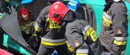 Na zdjęciu strażaków ubranych w ubrania bojowe wydostających poszkodowaną osobę (manekina) z samochodu osobowego, który leży na boku. Strażacy wcześniej usunęli dach pojazdu. W pobliżu leży deska ortopedyczna, na której będą wyciągać osobę poszkodowaną. W tyle widzimy zniszczone samochody na autozłomie ułożone jeden na drugim.